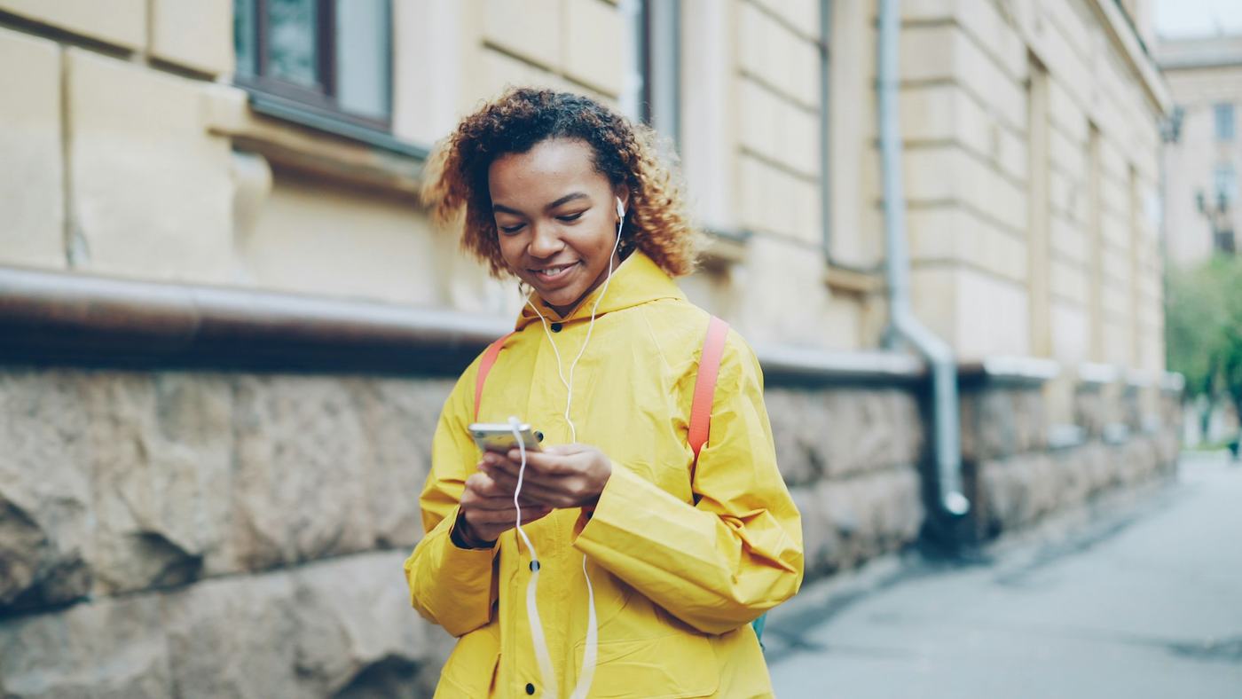 Eine junge Frau in gelber Jacke lacht auf ihr Smartphone.
