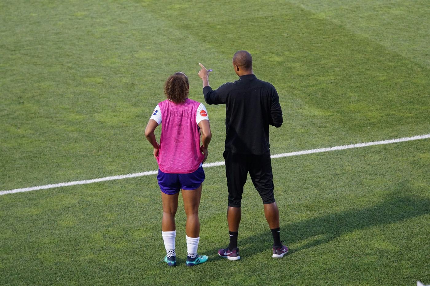 Ein Trainer und ein Kind stehen von hinten gesehen auf dem Fußballplatz.